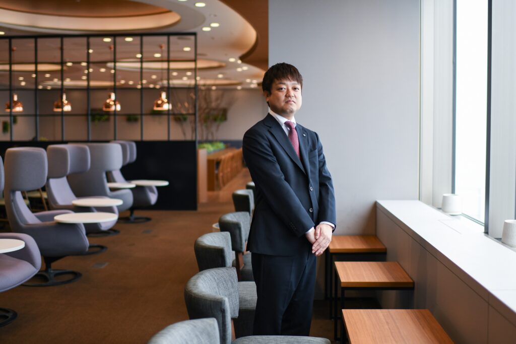 A man in a dark suit stands by a window.