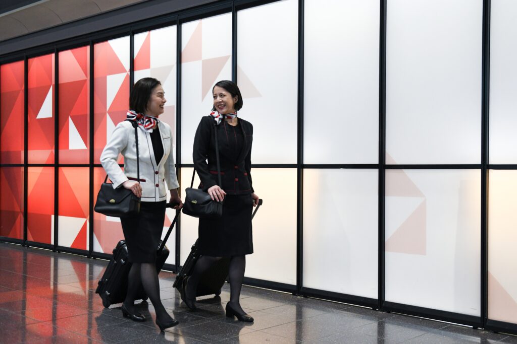 Two women in flight attendant uniforms chat with each other while walking with roller bags.