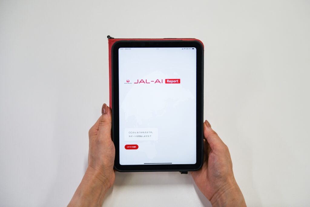 A tablet held in two hands with the JAL AI-Report app on screen.