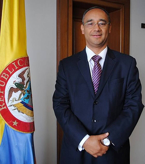A man in a suit stands next to the Colombian flag with his hands folded in front of him